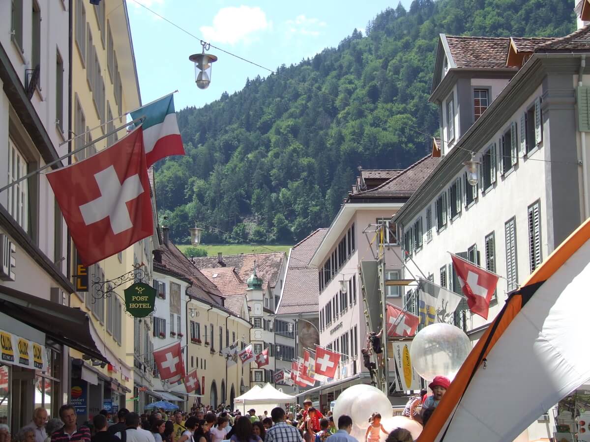 zdjęcie szwajcarskiego miasteczka, w tle góry, po lewej flaga szwjacarii